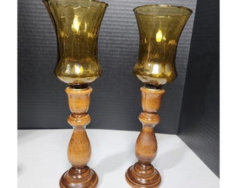 May include: Two vintage candle holders. Each has a dark amber glass shade with a textured rim, atop a turned wooden base. The wooden bases are a medium brown color, with a round base and a decorative stem. The candle holders are displayed on a white surface.