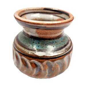 May include: A small, handcrafted ceramic vase with a unique design. The vase features a rounded body with a textured base and a flared rim. The color palette includes shades of brown, teal, black, and white, creating a visually appealing aesthetic. The vase is suitable for small flowers.