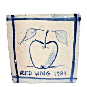 May include: Square ceramic piece with a cream-colored background and a blue apple design. The apple has two leaves and the text "RED WING 1984" in blue. A blue border frames the design.