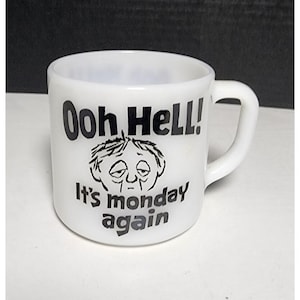 May include: White ceramic mug with a curved handle. The mug features the phrase "Ooh HeLL! It's monday again" in black text, along with a cartoon illustration of a tired face.