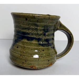 May include: Handmade ceramic mug with a unique design. The mug features a green and blue glaze with textured wave-like patterns. The handle is sturdy and the mug has a rustic, artisanal feel. Perfect for coffee or tea.