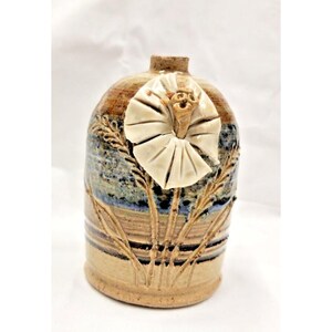 May include: Handmade ceramic vase with a beige, brown, and blue color scheme. The vase features a large, cream-colored flower and wheat stalks. The vase has a rounded shape and a textured surface, adding to its rustic charm.