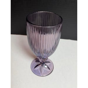 May include: A translucent purple glass goblet featuring a vertical ribbed pattern along the bowl. The stem connects to a circular base that displays a molded star-shaped design radiating from the center point of the foot of the drinking vessel.