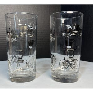 May include: Two clear drinking glasses with a black and white carriage design. The glasses are cylindrical and feature a repeating pattern of vintage carriages, adding a touch of classic charm to any table setting.