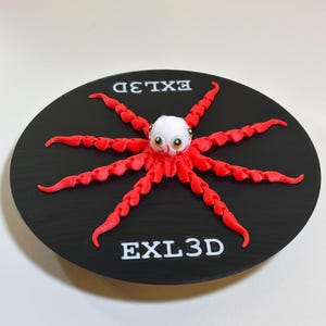 May include: A 3D-printed octopus figurine with red tentacles and a white head, set on a black circular base. The text "EXL3D" is printed in white on the base. The octopus has large, cartoonish eyes.