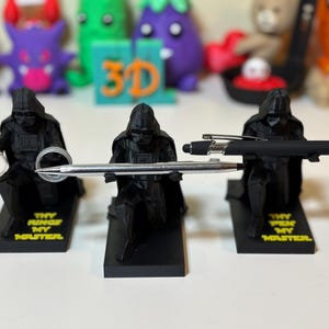 May include: Three black Darth Vader figurines holding a ring and pens. The figurines are on black bases with yellow text that reads "THY RINGS MY MASTER."