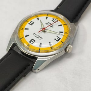 K&ouml;nnte beinhalten: Eine Vintage-Armbanduhr mit wei&szlig;em Zifferblatt, schwarzen Ziffern und einem gelben Au&szlig;enring. Die Uhr hat ein silbernes Geh&auml;use und ein schwarzes Lederarmband. Der Text "PARA SHOCK 17 JEWELS" ist sichtbar.