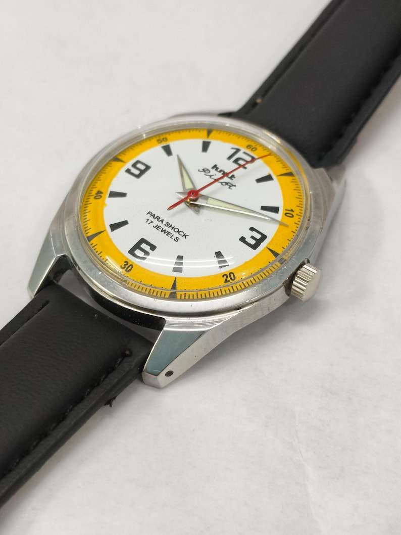 K&ouml;nnte beinhalten: Eine Vintage-Armbanduhr mit wei&szlig;em Zifferblatt, schwarzen Ziffern und einem gelben Au&szlig;enring. Die Uhr hat ein silbernes Geh&auml;use und ein schwarzes Lederarmband. Der Text "PARA SHOCK 17 JEWELS" ist sichtbar.