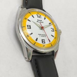 K&ouml;nnte beinhalten: Eine Vintage HMT Pilot Armbanduhr mit wei&szlig;em Zifferblatt, gelben Akzenten und schwarzem Lederarmband. Die Uhr hat ein silberfarbenes Geh&auml;use und einen roten Sekundenzeiger. Das Zifferblatt zeigt den Text "Pilot" und "Para Shock 17 Jewels".