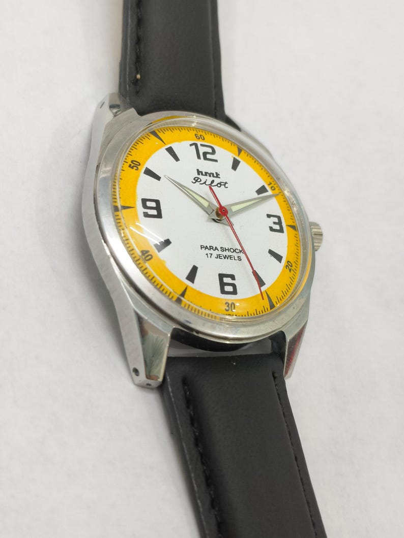 K&ouml;nnte beinhalten: Eine Vintage HMT Pilot Armbanduhr mit wei&szlig;em Zifferblatt, gelben Akzenten und schwarzem Lederarmband. Die Uhr hat ein silberfarbenes Geh&auml;use und einen roten Sekundenzeiger. Das Zifferblatt zeigt den Text "Pilot" und "Para Shock 17 Jewels".