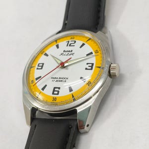 K&ouml;nnte beinhalten: Eine Vintage-Armbanduhr mit wei&szlig;em Zifferblatt und gelbem Au&szlig;enring. Die Uhr hat schwarze Ziffern, einen roten Sekundenzeiger und den Text "hmt Pilot" und "PARA SHOCK 17 JEWELS". Sie hat ein silbernes Geh&auml;use und ein schwarzes Lederarmband.