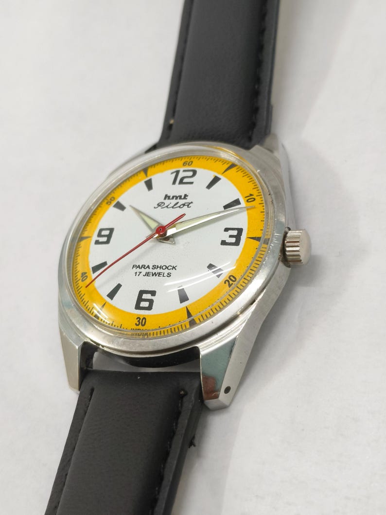 K&ouml;nnte beinhalten: Eine Vintage-Armbanduhr mit wei&szlig;em Zifferblatt und gelbem Au&szlig;enring. Die Uhr hat schwarze Ziffern, einen roten Sekundenzeiger und den Text "hmt Pilot" und "PARA SHOCK 17 JEWELS". Sie hat ein silbernes Geh&auml;use und ein schwarzes Lederarmband.