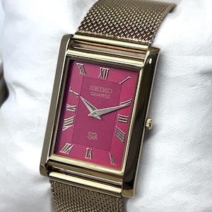 May include: A rectangular gold-tone watch with a vibrant magenta dial featuring a wavy line pattern. The watch face displays silver-tone Roman numeral hour markers, silver hands, and the text SEIKO QUARTZ. It has a gold-tone mesh metal strap.
