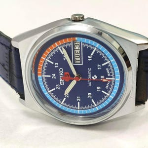 May include: A silver-toned Seiko 5 automatic watch with a blue dial and a blue leather strap. The watch face features orange and light blue accents, white numerals, and a day/date display. The watch is approximately 1.5 inches in diameter.