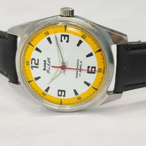 K&ouml;nnte beinhalten: Eine Vintage-Armbanduhr mit silberfarbenem Geh&auml;use und schwarzem Lederarmband. Das Zifferblatt ist wei&szlig; mit schwarzen Ziffern und einem gelben Au&szlig;enring. Die Zeiger sind silber und rot. Der Text "hmt Pilot" und "Para Shock 17 Jewels".