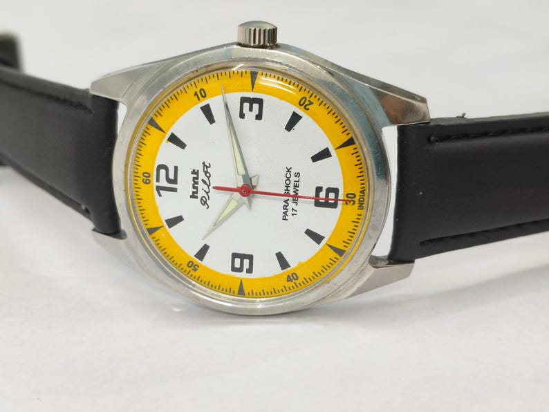 K&ouml;nnte beinhalten: Eine Vintage-Armbanduhr mit silberfarbenem Geh&auml;use und schwarzem Lederarmband. Das Zifferblatt ist wei&szlig; mit schwarzen Ziffern und einem gelben Au&szlig;enring. Die Zeiger sind silber und rot. Der Text "hmt Pilot" und "Para Shock 17 Jewels".
