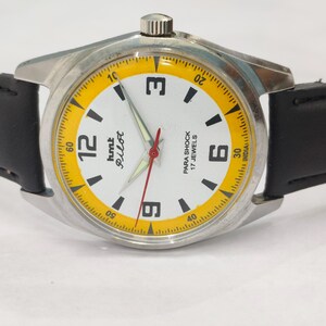 K&ouml;nnte beinhalten: Silberfarbene Armbanduhr mit wei&szlig;em Zifferblatt, schwarzen Zeigern und einem roten Sekundenzeiger. Die Uhr hat einen gelben Au&szlig;enring mit schwarzen Markierungen und ein schwarzes Lederarmband. Das Zifferblatt zeigt "hmt Pilot" und "PARA SHOCK 17 JEWELS".