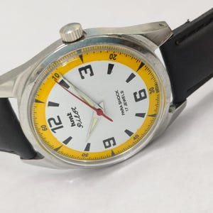 K&ouml;nnte beinhalten: Eine silberfarbene Armbanduhr mit wei&szlig;em Zifferblatt und schwarzem Lederarmband. Das Zifferblatt hat schwarze Ziffern, einen gelben Au&szlig;enring und den Text "hmt Pilot". Die Zeiger der Uhr sind rot und silber.