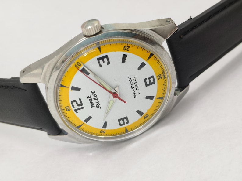 K&ouml;nnte beinhalten: Eine silberfarbene Armbanduhr mit wei&szlig;em Zifferblatt und schwarzem Lederarmband. Das Zifferblatt hat schwarze Ziffern, einen gelben Au&szlig;enring und den Text "hmt Pilot". Die Zeiger der Uhr sind rot und silber.