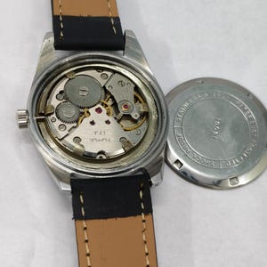 K&ouml;nnte beinhalten: Eine Vintage-Armbanduhr mit silberfarbenem Geh&auml;use und braunem Lederarmband. Das Innenleben der Uhr ist sichtbar, mit Zahnr&auml;dern und kleinen roten Juwelen. Der Geh&auml;useboden ist abgenommen und zeigt die Worte "Stainless Steel" und "Shock Proof".
