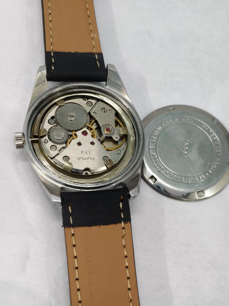 K&ouml;nnte beinhalten: Eine Vintage-Armbanduhr mit silberfarbenem Geh&auml;use und braunem Lederarmband. Das Innenleben der Uhr ist sichtbar, mit Zahnr&auml;dern und kleinen roten Juwelen. Der Geh&auml;useboden ist abgenommen und zeigt die Worte "Stainless Steel" und "Shock Proof".