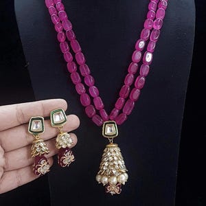 Op de afbeelding: Een magenta kralenketting met bijpassende oorbellen. De ketting heeft een hanger in de vorm van een goudkleurige bel met witte parels. De oorbellen hebben een vierkante groene en witte bovenkant met een belvorm en magenta accenten.
