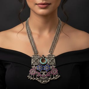 May include: A silver statement necklace with a rectangular pendant featuring turquoise, blue, and pink accents. The necklace has a detailed design and is worn against a black top. The model has dark hair and is looking at the camera.