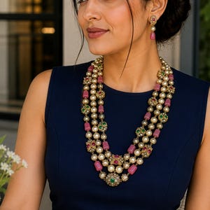 May include: A woman wearing a layered necklace with pink, green, and white gemstones. The necklace is paired with matching earrings. She is wearing a sleeveless navy blue dress. A cup of coffee sits on a table in the foreground.