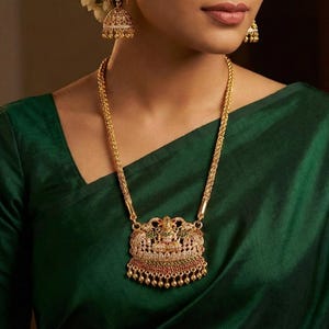 Könnte beinhalten: Ein goldenes Halsketten- und Ohrring-Set. Die Halskette hat einen detaillierten Anhänger mit aufwendigen Designs und farbigen Edelsteinen. Die Ohrringe sind glockenförmig mit ähnlichen Details. Der Schmuck wird mit einem grünen Oberteil getragen.