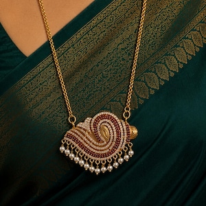 May include: A gold-toned necklace with a pendant featuring a swirling design of red and clear stones, accented with small white pearls. The necklace hangs against a dark green background.