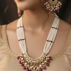 Op de afbeelding: Een gouden ketting en bijpassende oorbellen set. De ketting heeft meerdere strengen witte kralen, geaccentueerd met rode kralen. De hanger heeft een gouden filigraan ontwerp met rode en witte accenten. De oorbellen hebben een vergelijkbaar ontwerp.