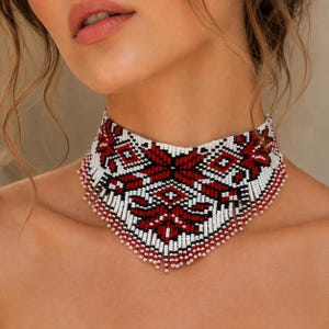 May include: A wide choker necklace crafted from small seed beads in a geometric pattern. The design features a repeating motif in deep red, black, and white, with a fringe of dangling beads along the bottom edge that follows a V-shaped contour.