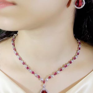 May include: A silver necklace and matching earrings set featuring teardrop-shaped red gemstones and small white stones. The necklace has a pendant with a large teardrop-shaped red stone. The earrings have a similar design.