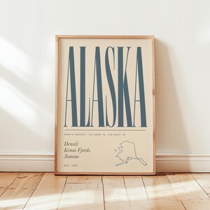 May include: A vertical rectangular wall art print on a light beige background featuring the word ALASKA in large blue sans-serif letters. Below the text are the coordinates 64.2008 N, 149.4937 W, the words Denali, Kenai Fjords, Juneau, and a map outline of Alaska.
