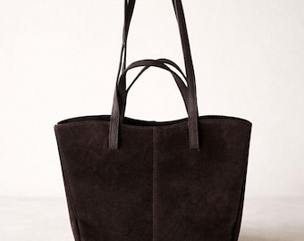May include: A dark brown suede tote bag with two long handles and two shorter handles. The bag has a rectangular shape with a flat bottom and a simple, minimalist design. The handles are a darker shade of brown.