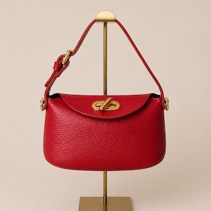 May include: A vibrant red handbag with a textured finish, featuring a flap closure and a gold-toned clasp. The bag has a top handle and is displayed on a gold-colored stand. The bag's design is classic and elegant.