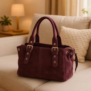 May include: A burgundy suede handbag with two top handles and gold-colored hardware. The bag features a rectangular shape with a zippered top and adjustable side straps. The bag is sitting on a beige sofa.