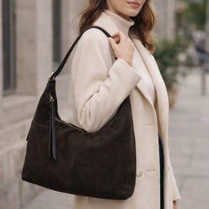 May include: A dark brown suede shoulder bag with a long strap and gold-colored zipper. The bag has a tassel and is being carried over the shoulder. The bag is a stylish accessory.