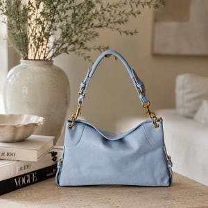 May include: A light blue shoulder bag with a textured surface, featuring gold-toned metal hardware, adjustable side straps with buckles, and a single handle attached to the bag with gold-toned lobster claw clasps on both sides.