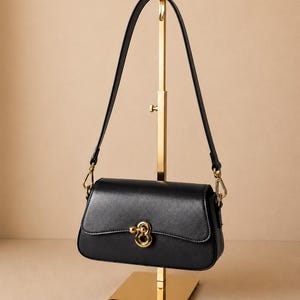 May include: A black rectangular shoulder bag with a smooth finish, featuring a gold-toned metal clasp on the front flap. The bag has a single thin black strap attached with gold-toned hardware clips on each side, hanging from a gold display stand.