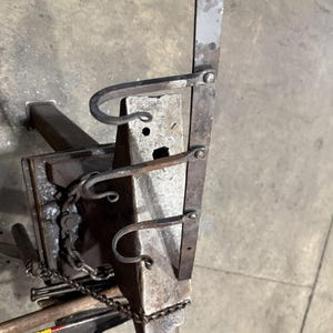以下が含まれることがあります： 手作りの金属製の金床で、先端が尖っており、曲がったフックが付いた垂直な金属片が特徴です。 金床は金属製のベースにセットされており、チェーンとツールが見えます。 金属は素朴で工業的な外観をしています。