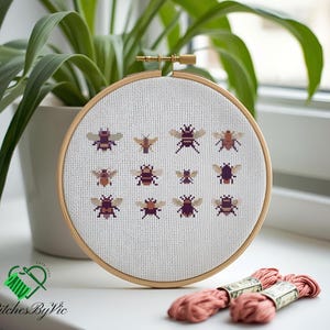 May include: A cross-stitch embroidery hoop featuring a pattern of pixelated bees in shades of brown, purple, and tan. The embroidery is set against a white fabric background, held in a wooden hoop. The text "Stitches By Vic" is visible.