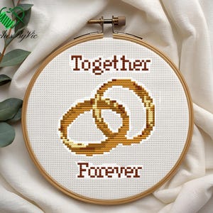 Op de afbeelding: Kruissteekborduurwerk in een houten hoepel. Het ontwerp toont twee in elkaar grijpende ringen in bruintinten, met de woorden "Together" en "Forever" boven en onder de ringen geborduurd. De achtergrond is wit.