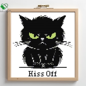 Op de afbeelding: Kruissteekkunst met een chagrijnige zwarte kat met felgroene ogen. De kat staat tegen een witte achtergrond, omlijst door licht hout. De tekst "Hiss Off" staat eronder.
