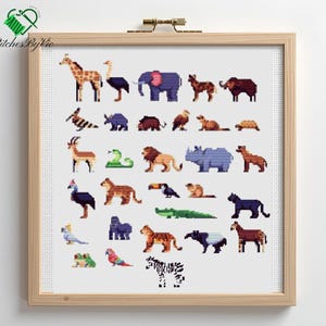 Op de afbeelding: Een ingelijste kruissteek met een verscheidenheid aan gepixelde dieren, waaronder een giraffe, een olifant, een leeuw en een zebra. Het kunstwerk is geplaatst tegen een witte achtergrond en ingelijst in een licht houten frame. De tekst "Stitches by Vie" staat linksboven.