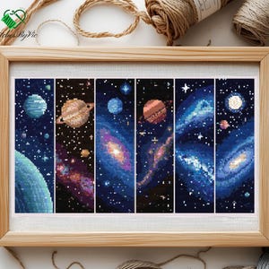 May include: A framed cross-stitch artwork depicting space. The design features planets, galaxies, and stars in shades of blue, purple, and orange. The artwork is presented in a light brown wooden frame.