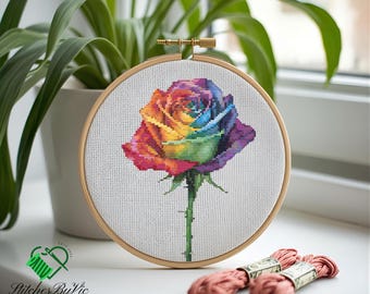 May include: A cross-stitch embroidery of a rainbow rose in a wooden hoop. The rose features vibrant colors including red, orange, yellow, green, blue, and purple. The stem is green, and the background is white. The brand name "Stitches By Vic" is visible.