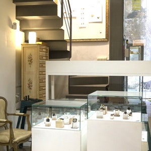 May include: A jewelry shop interior with white display cases showcasing jewelry and watches. The shop has a white counter, a wooden cabinet, and a staircase leading to an upper level.
