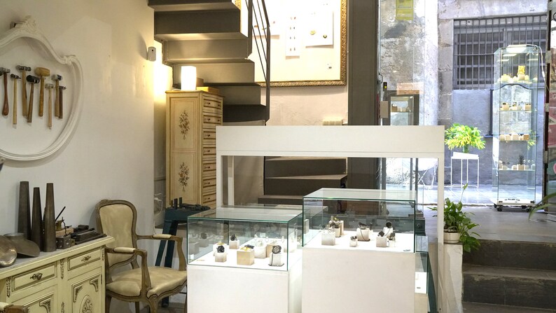 May include: A jewelry shop interior with white display cases showcasing jewelry and watches. The shop has a white counter, a wooden cabinet, and a staircase leading to an upper level.