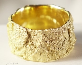 May include: A thick gold band ring with a textured, rough surface. The ring is made of solid gold and has a wide band.